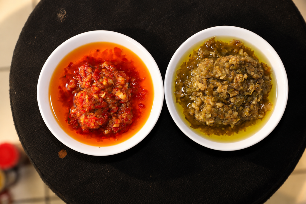 Dua piring kecil berisi sambal bawang cabai rawit merah dan hijau di atas meja hitam.